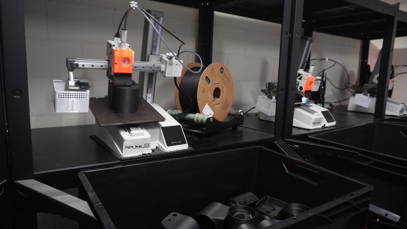 A close-up of a single Bambu Lab A1 mini printer labelled FARM_MINI_03 on a rack, with a bin underneath already half-full of finished black 3D-printed parts