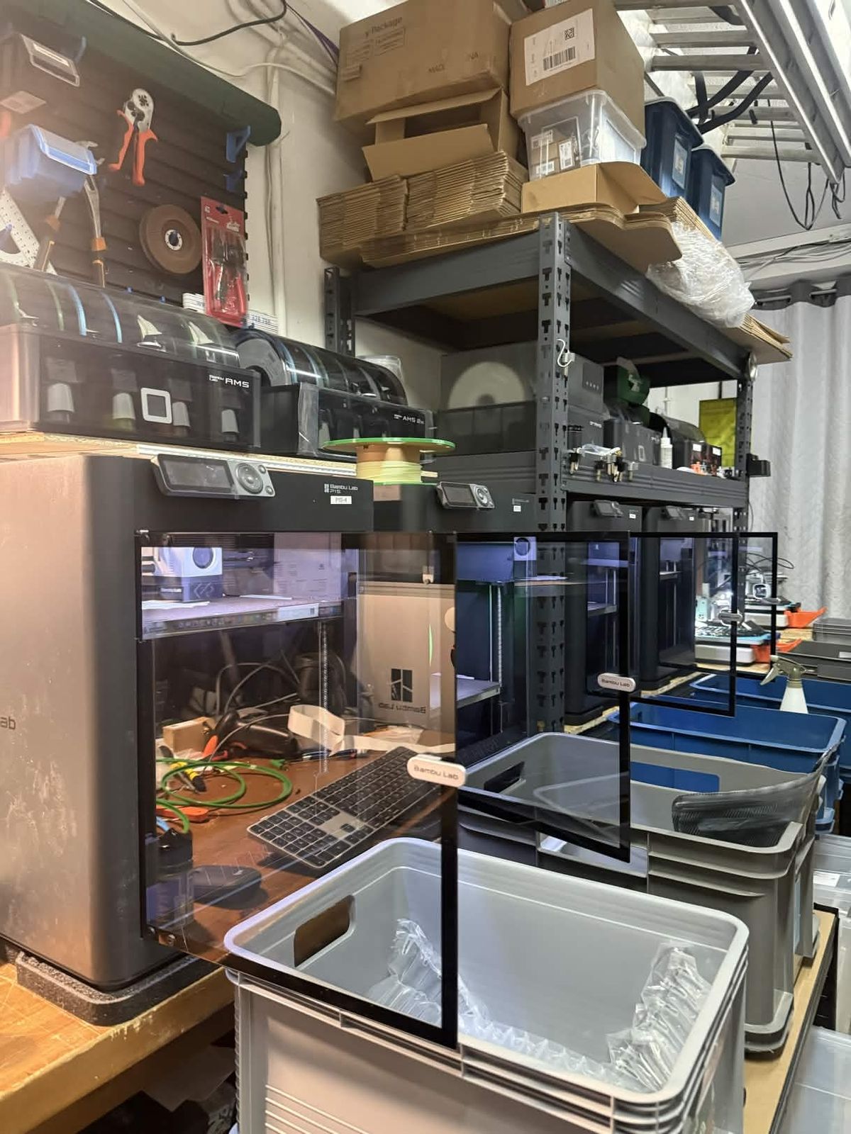 Several enclosed Bambu Lab printers with stacked AMS units on industrial shelving over Patryk's workbench, with tools, cabling and shipping totes