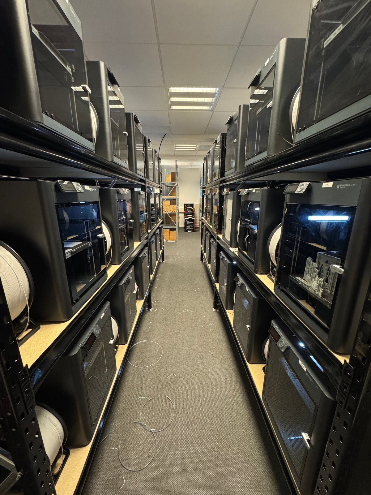 Aisle down two long racks packed with enclosed Bambu Lab printers and filament spools in LAB3D's farm