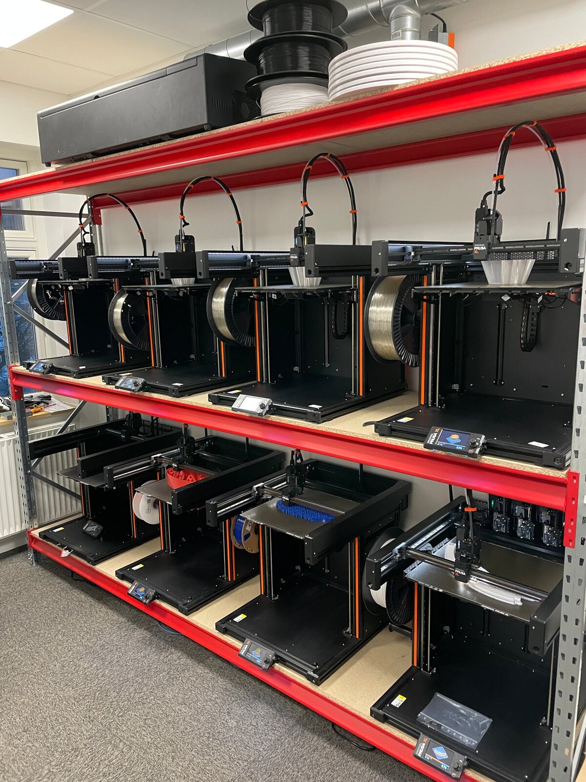 A red industrial rack with two shelves of Original Prusa printers and filament spools in LAB3D's farm
