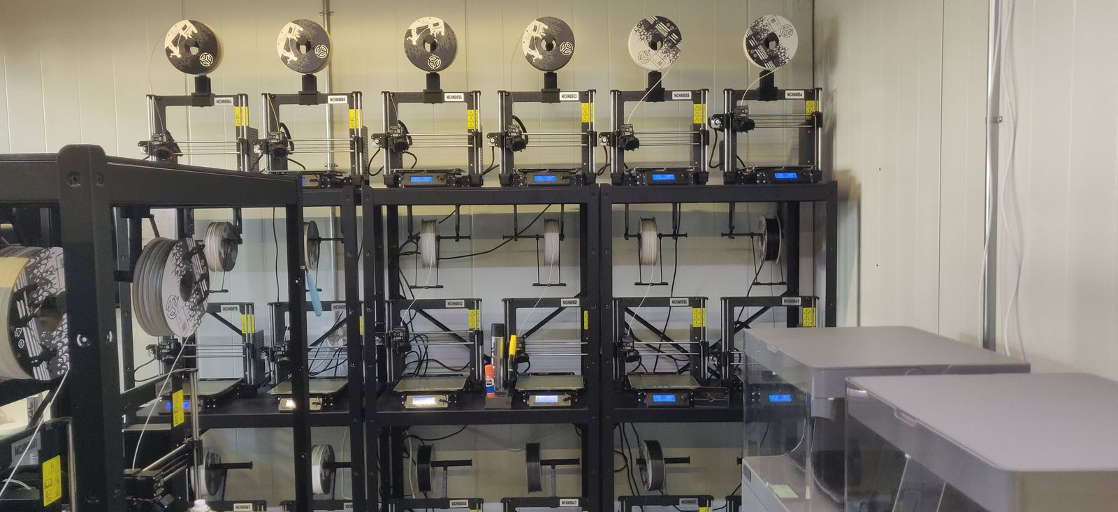 A multi-tier black rack of Prusa printers with branded spools feeding from above and blue control screens