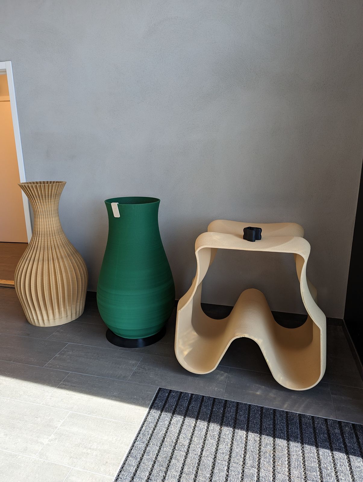 Two large 3D-printed floor vases and a sculpted stool against a grey wall