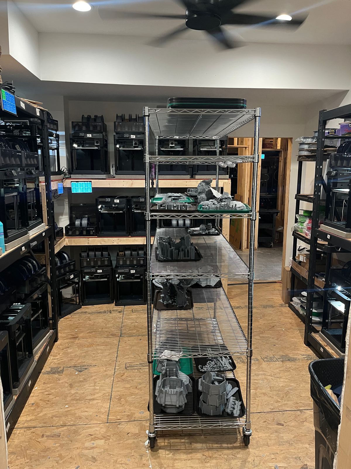 A wire shelving unit displaying finished grey terrain and scenery miniatures, flanked by racks of Bambu Lab P1 printers