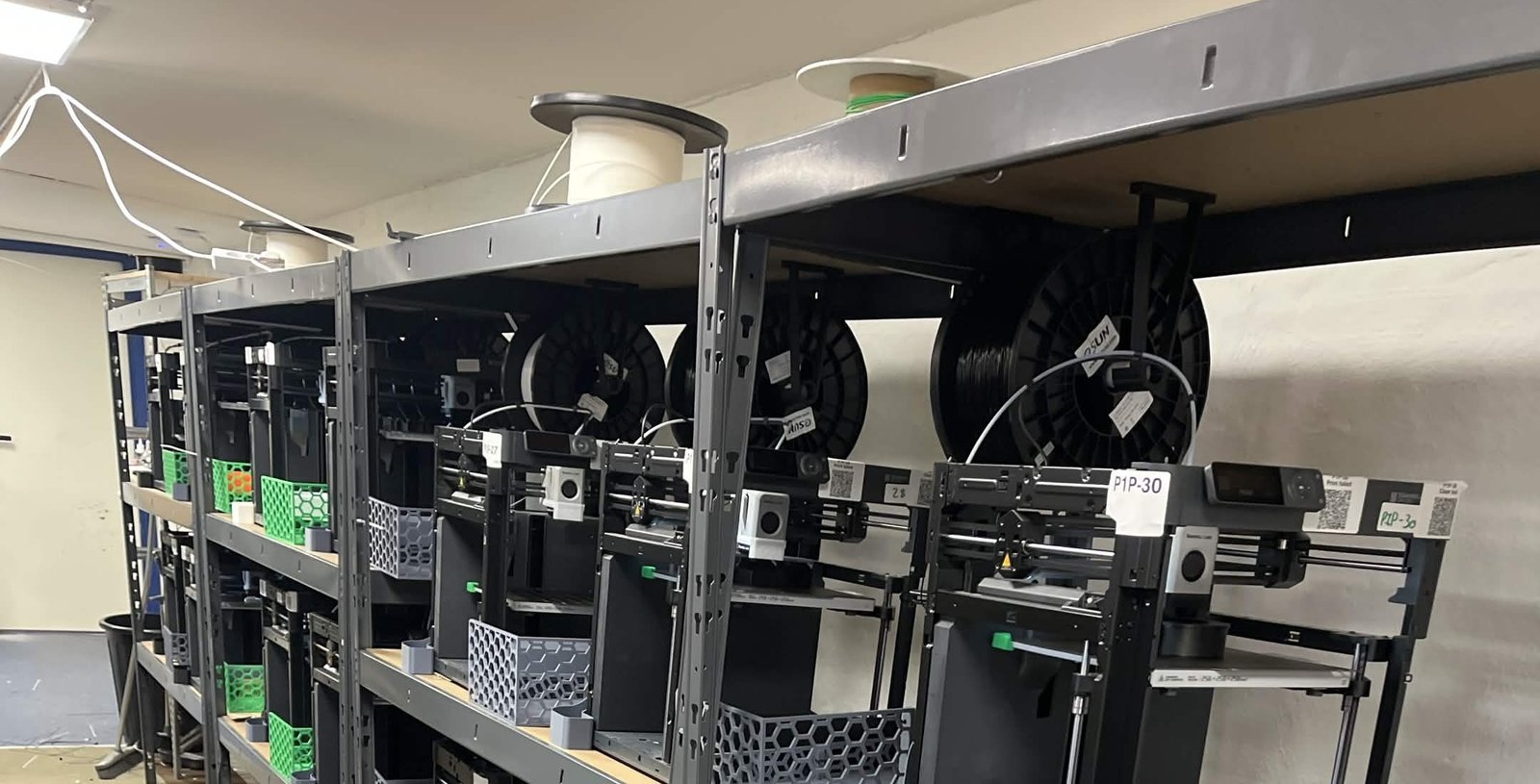 A long row of black Bambu Lab P1P printers on steel shelving at Weshape, filament spools mounted on top, one labelled P1P-30
