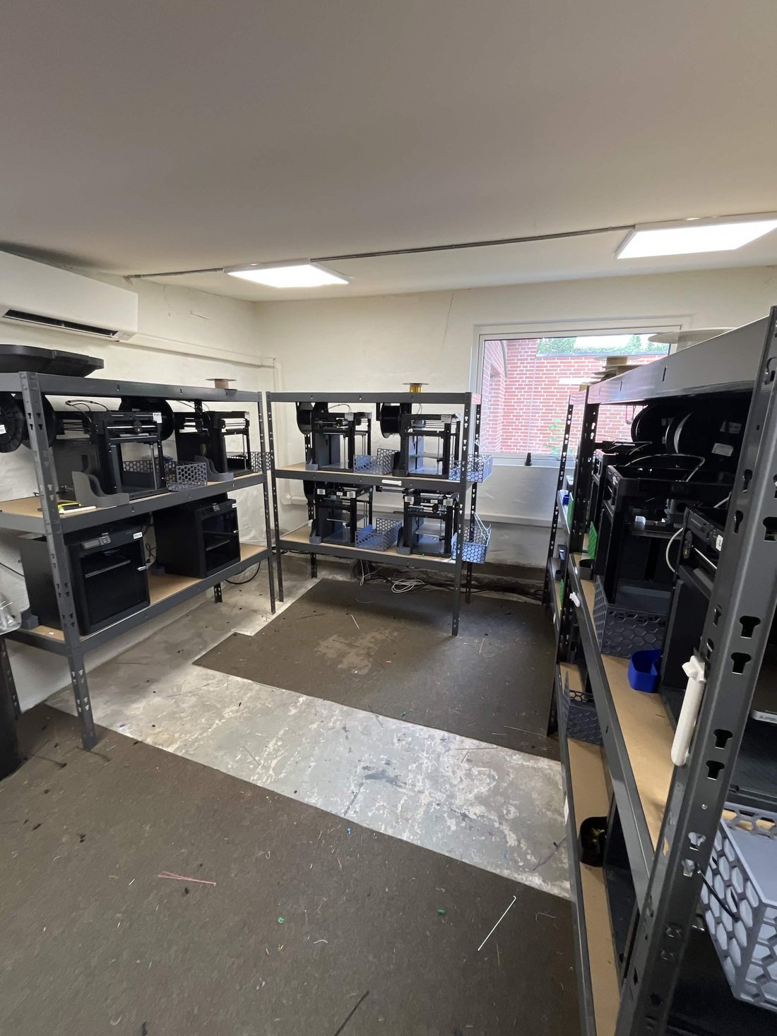Weshape's print-farm room: grey steel shelving racks of black Bambu Lab P1P printers in a clean white-walled space