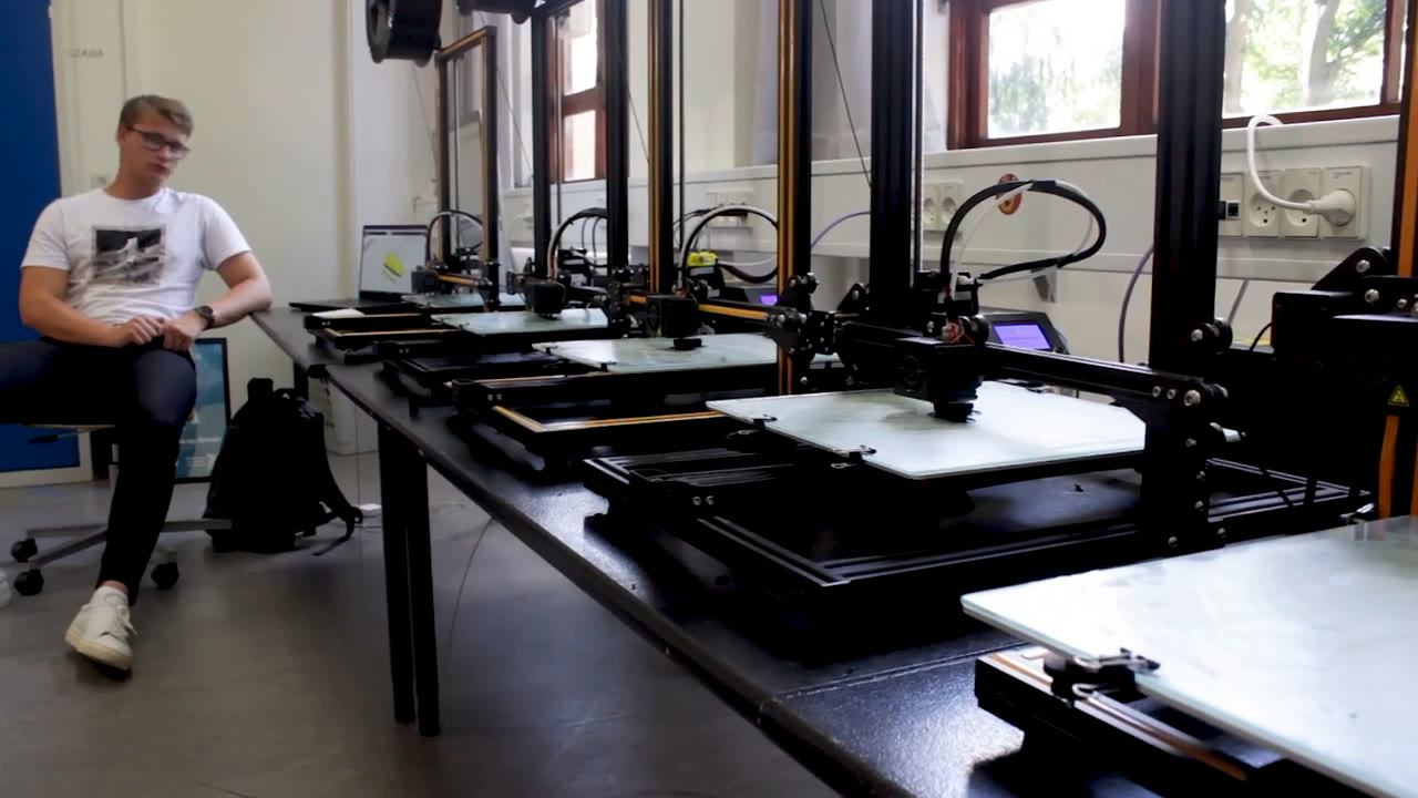 A long workshop table lined with open-frame 3D printers at the Media High School of Copenhagen, a student seated at the far end