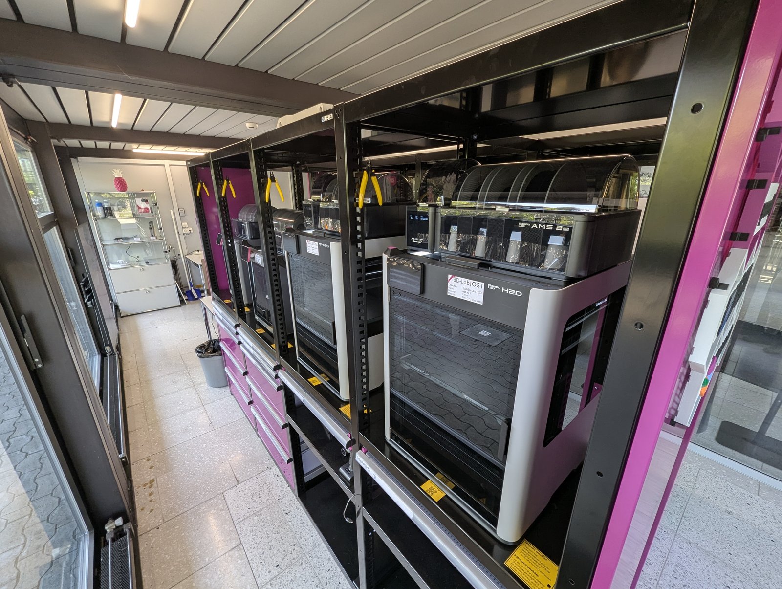 A row of enclosed Bambu Lab printers with AMS units on the magenta rack in the OST 3D-Lab