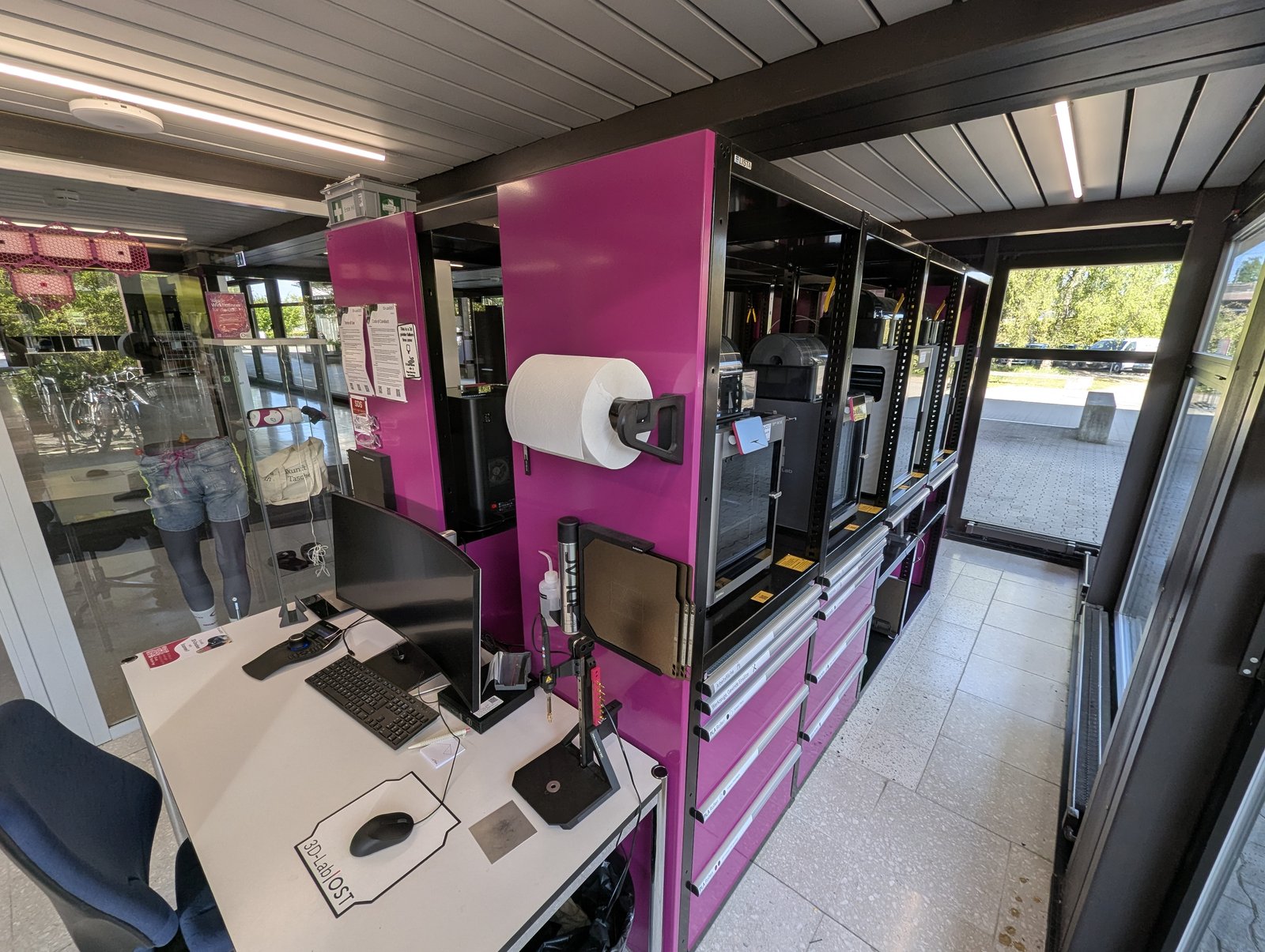 Side view of the OST 3D-Lab: the operator desk with monitors on the left, the magenta Bambu Lab printer rack on the right