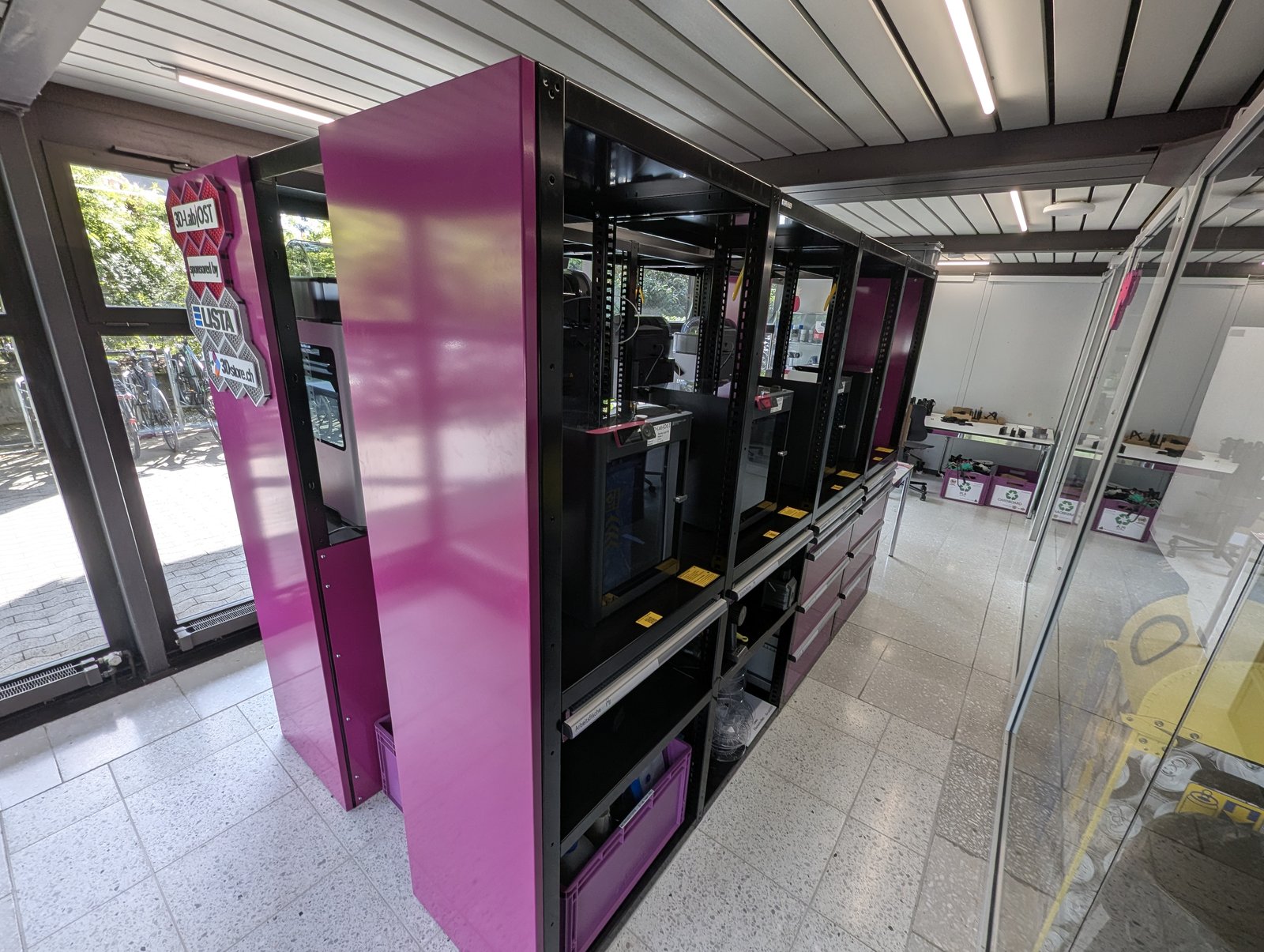 The OST 3D-Lab printer rack in magenta cabinets, glass walls and a lakeside-town view behind, finished prints on a shelf