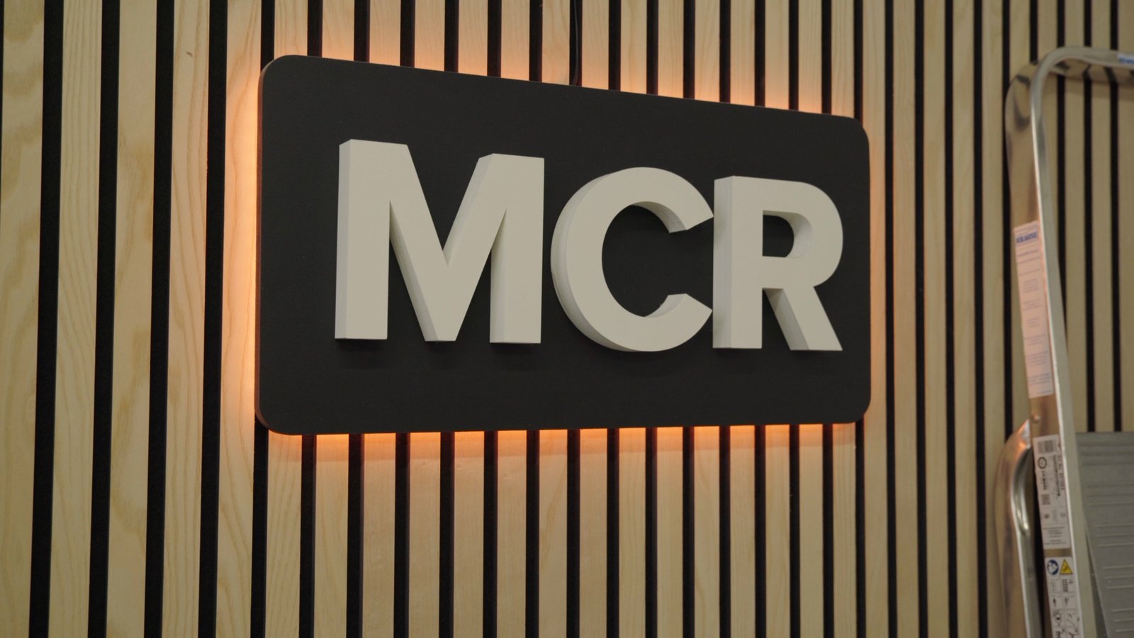 Large 3D-printed white MCR letters on a black backlit plate mounted on a wood-slat acoustic wall