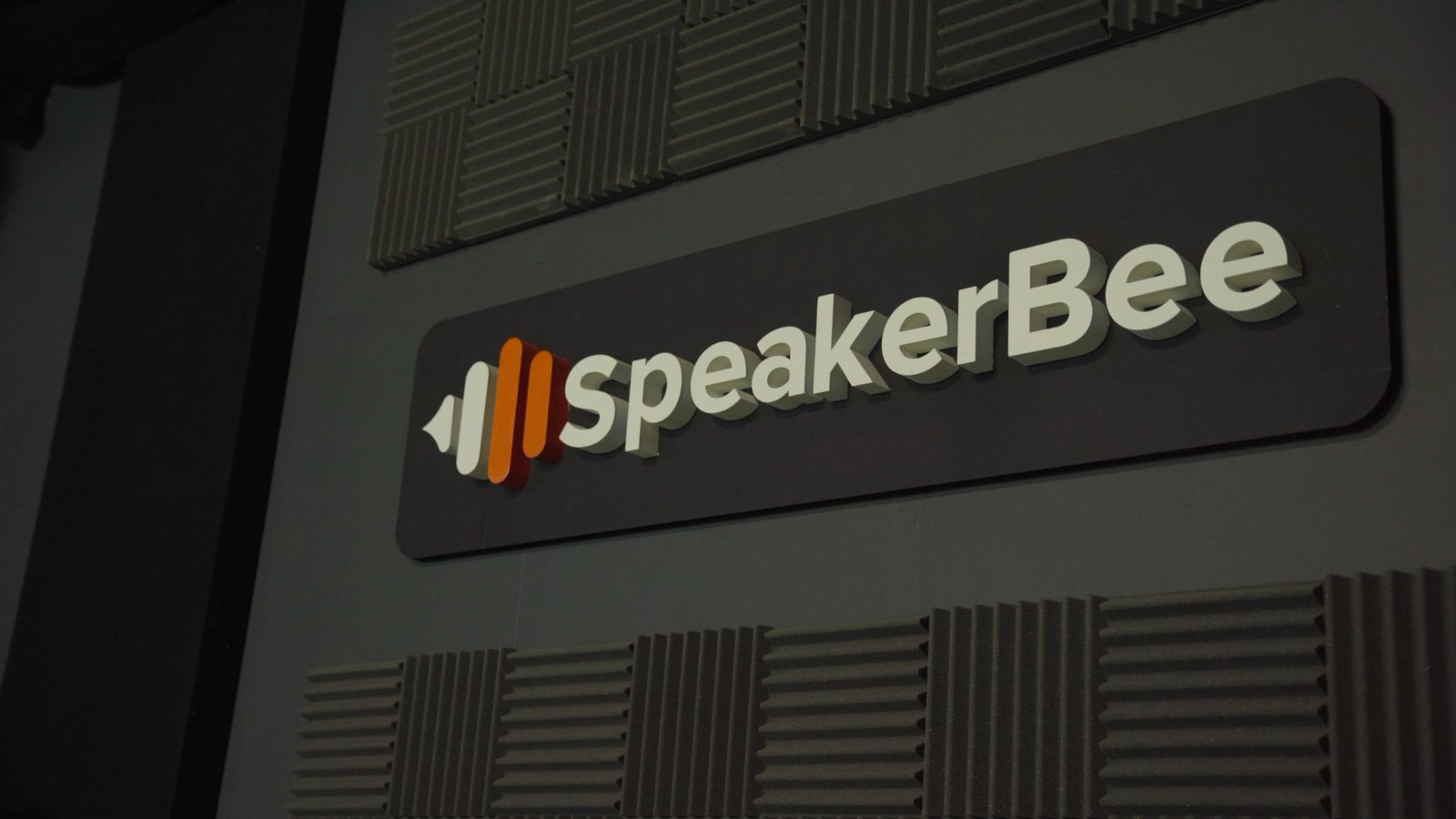 A 3D-printed SpeakerBee logo sign with white lettering and an orange sound-wave icon mounted between dark acoustic foam panels