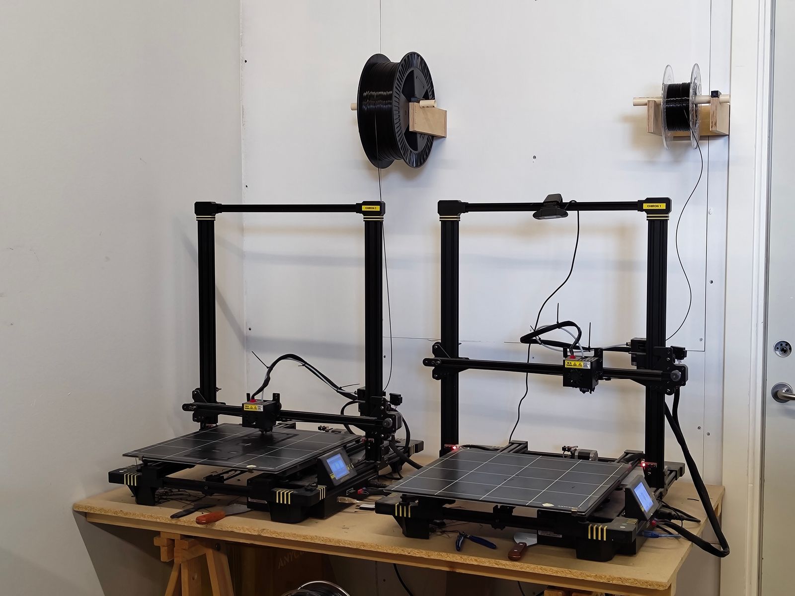 Two Anycubic Chiron 3D printers side by side on a wooden studio workbench with wall-mounted spool holders feeding filament from above